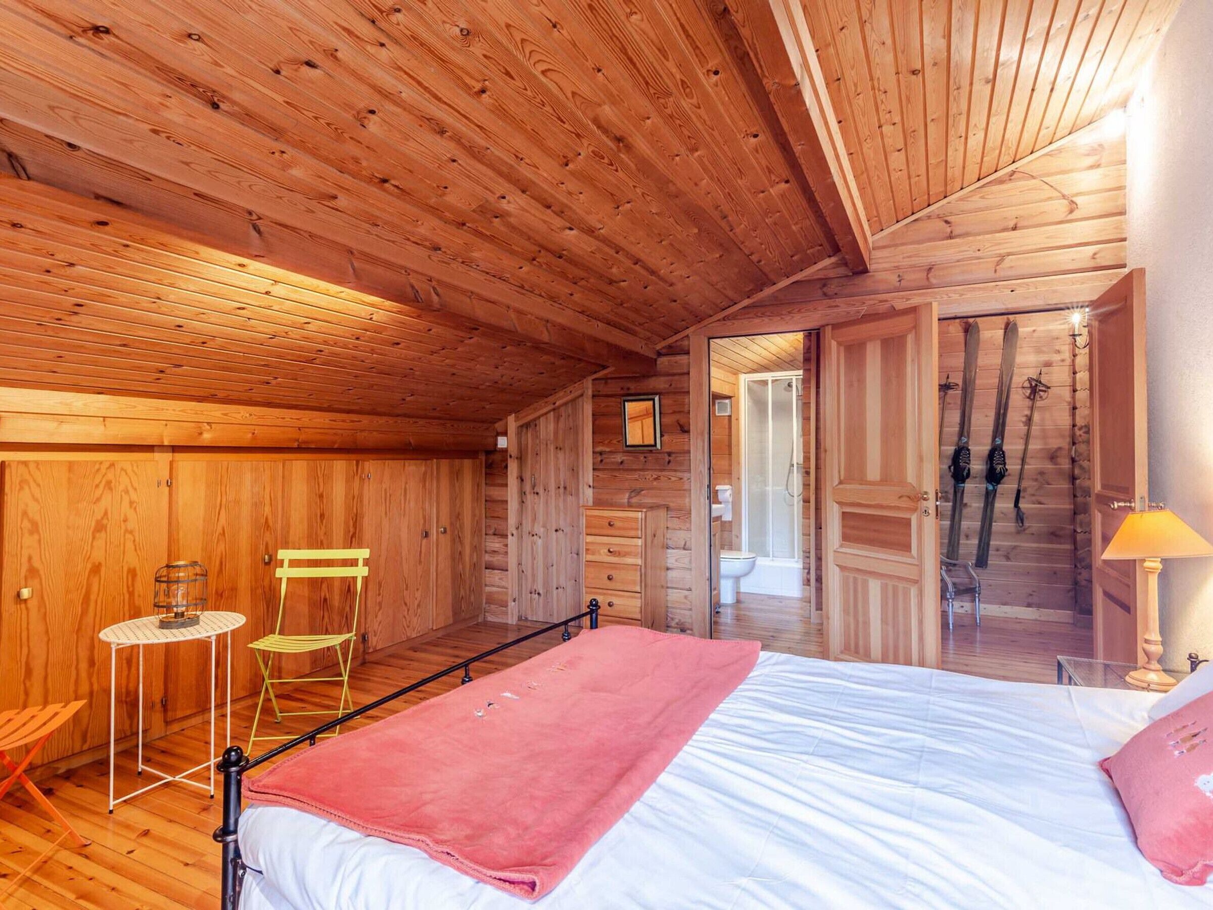 Une chambre à coucher située dans une cabane en bois, avec un lit, une chaise, une petite table et une salle de bain visible à travers une porte ouverte.