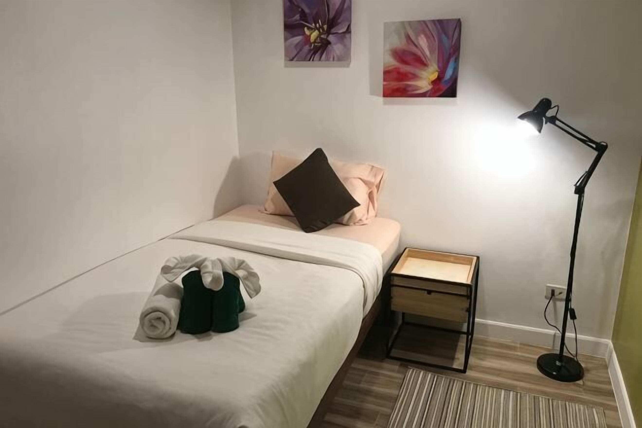A single bed with a white bedspread, a small bedside table, a lamp, and two wall-mounted paintings.