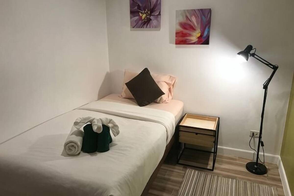 A single bed with a white bedspread, a small bedside table, a lamp, and two wall-mounted paintings.