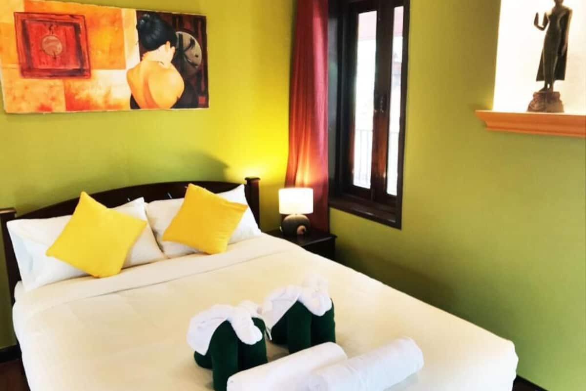 A hotel room with a bed, pillows, a nightstand, a lamp, a window with curtains, and a statue on a shelf.