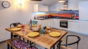 Une cuisine moderne avec une table à manger en bois pour quatre personnes, un four encastré et un tableau abstrait aux couleurs vives accroché au-dessus du plan de travail.
