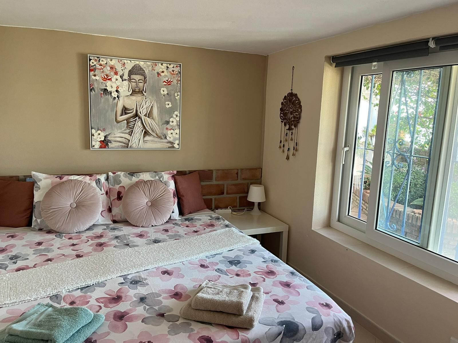 A bedroom with a bed, pillows, a bedside table, a lamp, a window, and a wall decoration.
