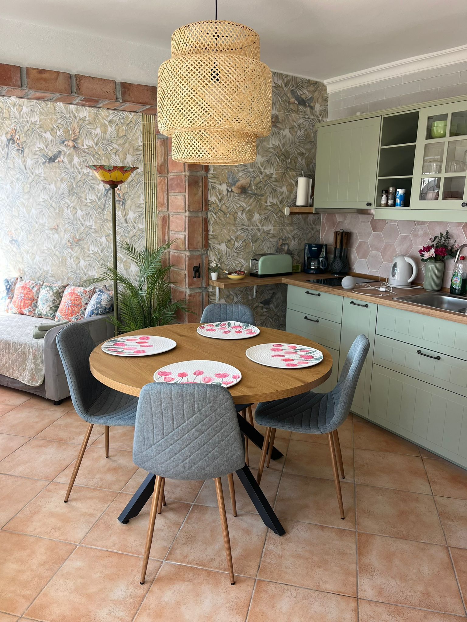 Un comedor con una mesa redonda, sillas tapizadas en gris, una lámpara colgante de ratán y una cocina al fondo.