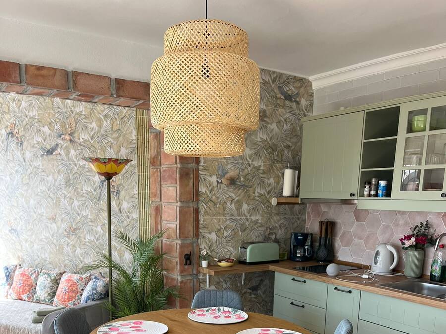 Un comedor con una mesa redonda, sillas tapizadas en gris, una lámpara colgante de ratán y una cocina al fondo.