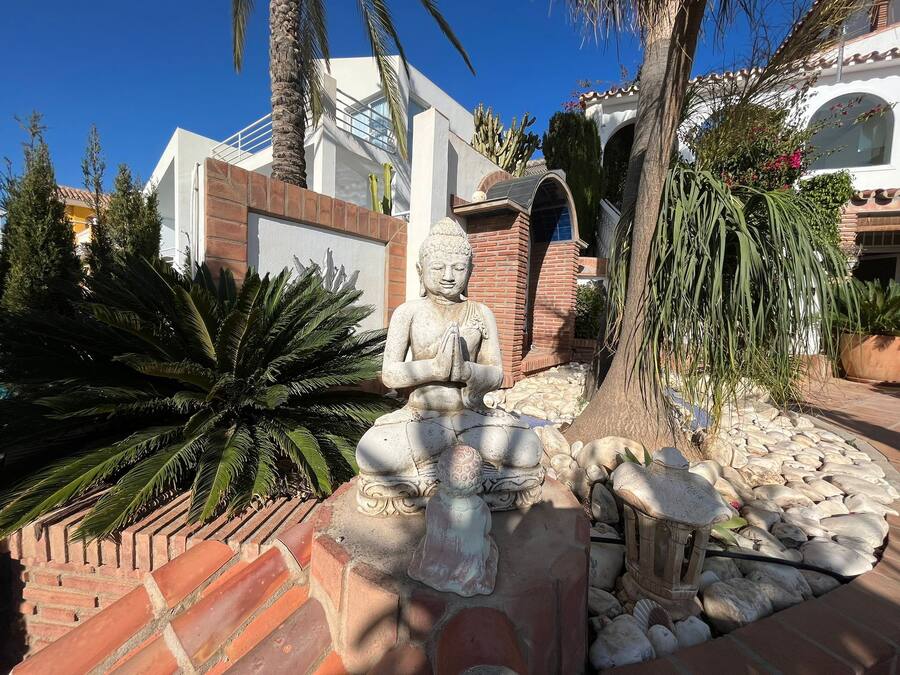 Una estatua de Buda rodeada de rocas y plantas, con un edificio moderno al fondo.