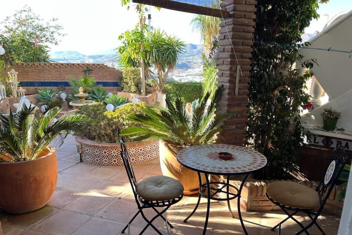Un patio al aire libre con una mesa redonda y dos sillas, rodeado de plantas en macetas y vistas de la ciudad.
