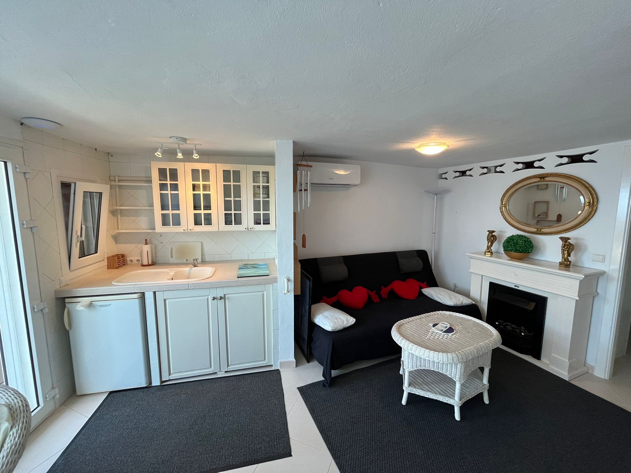 Un espacio habitable compacto con kitchenette, un sofá negro con cojines rojos, una pequeña mesa redonda y una chimenea.