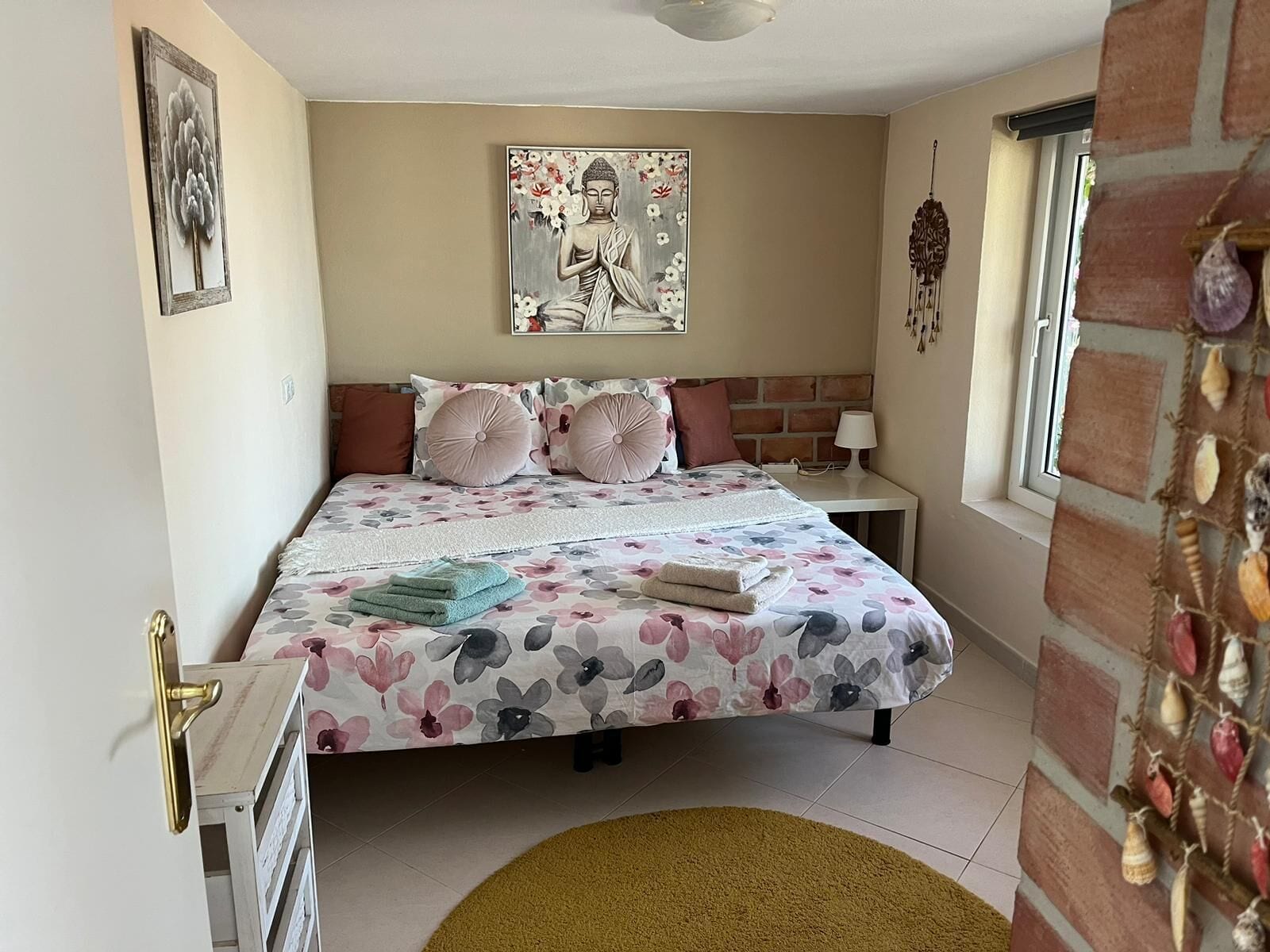 Un dormitorio con cama, almohadas, mesita de noche, lámpara, un cuadro y una ventana.