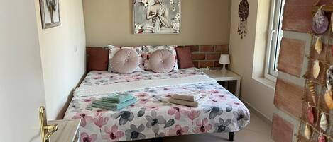 Un dormitorio con cama, almohadas, mesita de noche, lámpara, un cuadro y una ventana.