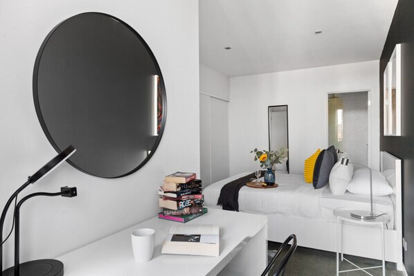 Una habitación de hotel moderna con un espejo grande y redondo, una mesita de noche con libros, un pequeño jarrón con flores y una cama con ropa de cama blanca y almohadones decorativos.