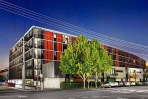 Un edificio moderno con fachada de vidrio y metal, un árbol en primer plano y coches aparcados a lo largo de la calle.
