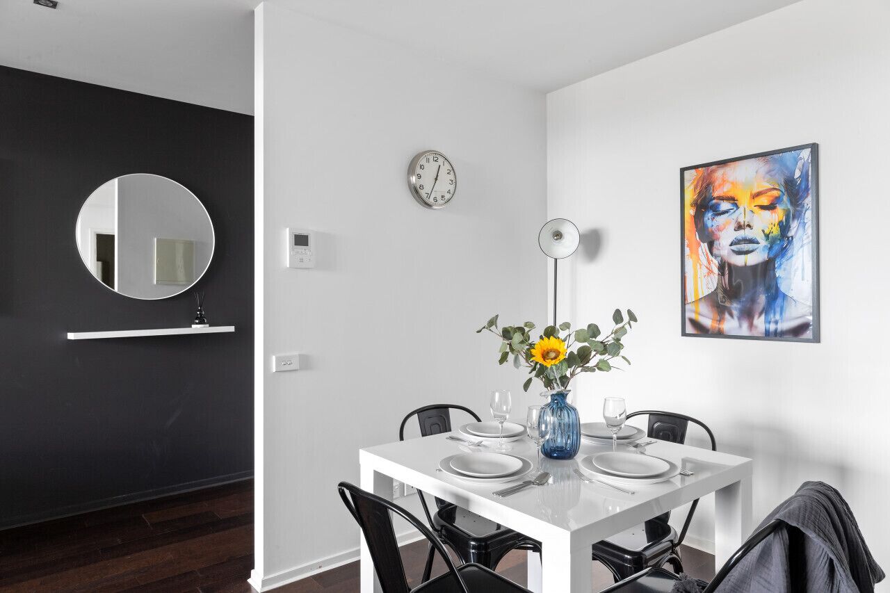 A modern dining area with a white table set for four, a round mirror, a wall clock, and a colorful abstract painting.