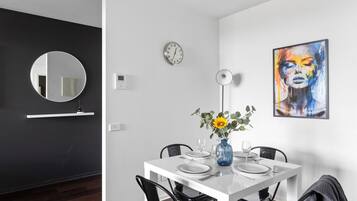 A modern dining area with a white table set for four, a round mirror, a wall clock, and a colorful abstract painting.