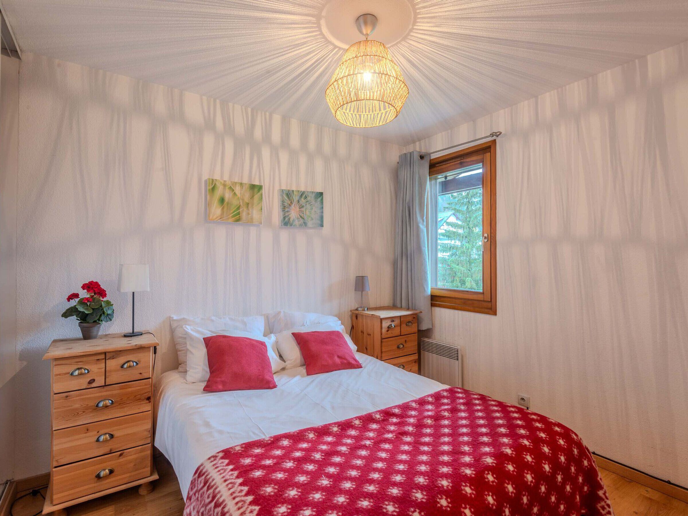 Un dormitorio con cama, cómoda de madera, manta roja estampada, dos obras de arte en la pared, ventana con vistas y lámpara colgante.