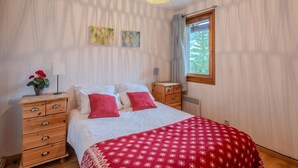 Un dormitorio con cama, cómoda de madera, manta roja estampada, dos obras de arte en la pared, ventana con vistas y lámpara colgante.