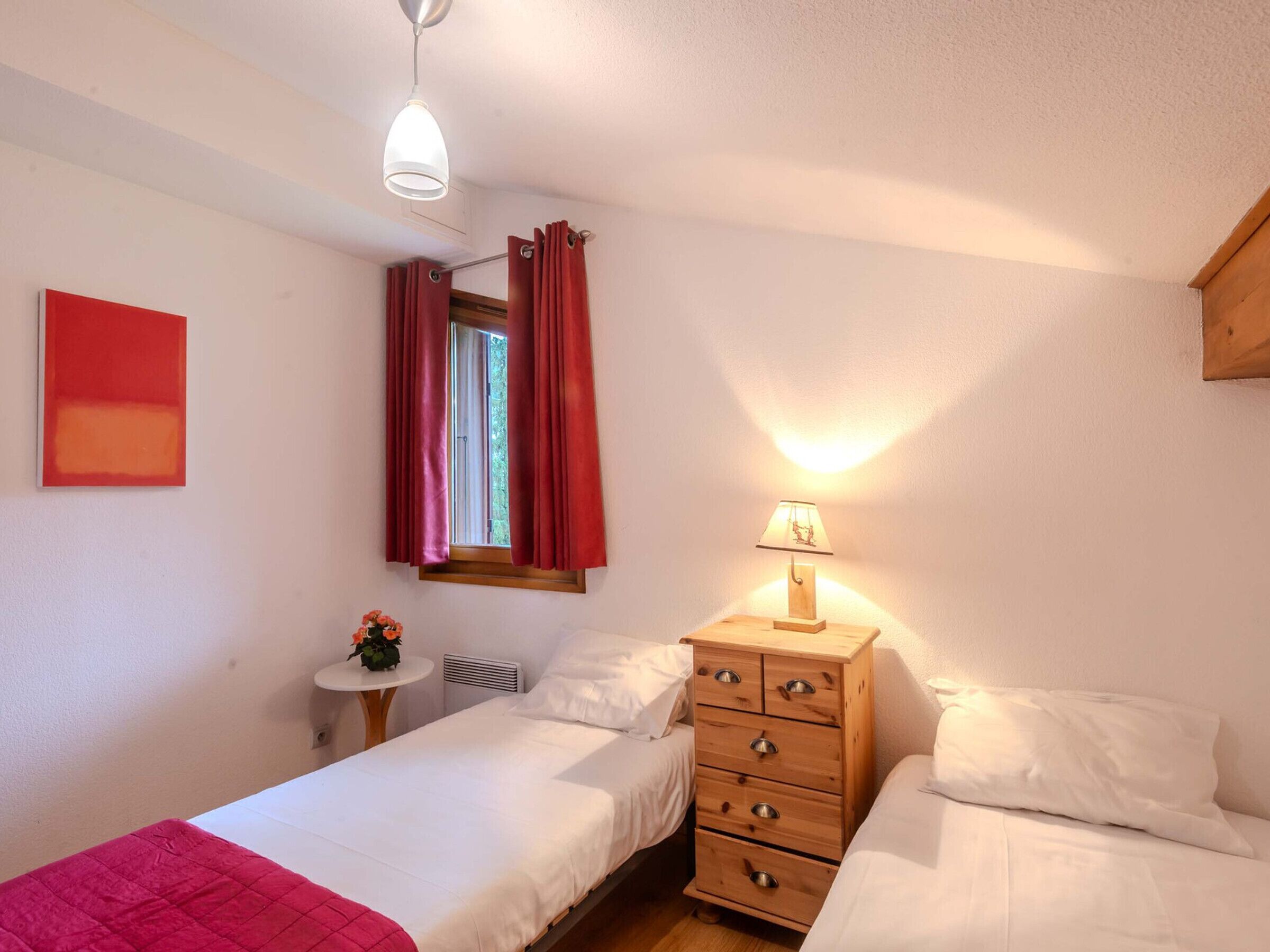 Een slaapkamer met twee bedden, een houten dressoir, een nachtkastje met een lamp en een raam met rode gordijnen.