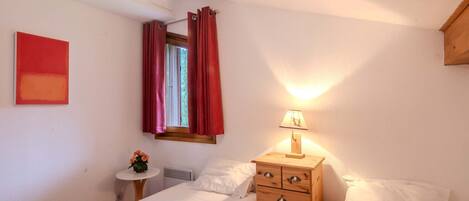 Een slaapkamer met twee bedden, een houten dressoir, een nachtkastje met een lamp en een raam met rode gordijnen.