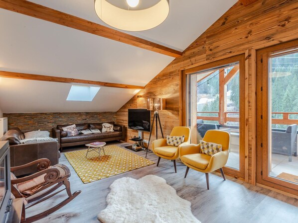 Una sala de estar estilo loft acogedora con pared de piedra, vigas de madera, un ventanal grande y una televisión.