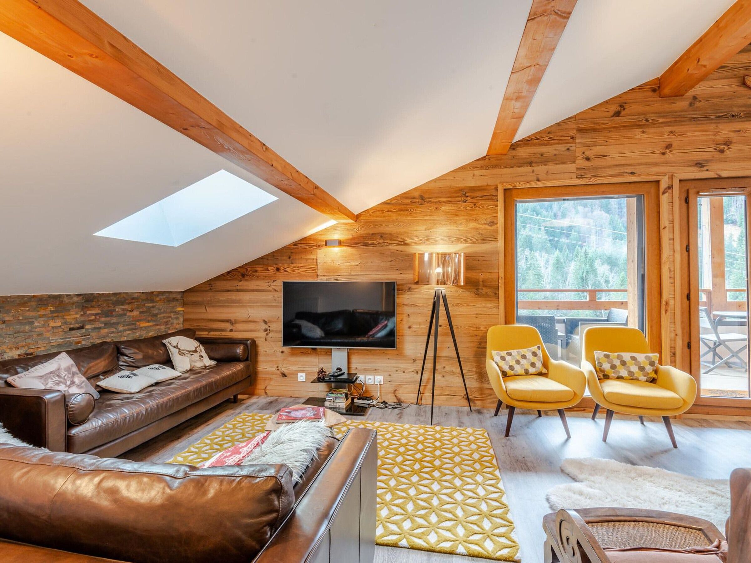 Een gezellige woonkamer met houten lambrisering, een schuin dak, een leren bank, gele fauteuils en een televisie.
