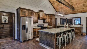 A spacious kitchen with wooden cabinets, a large island with bar stools, a stainless steel refrigerator, and a hanging light fixture. - Sweet Magnolia~Sleeps 10~Game Area~2 Masters (Broken Bow)