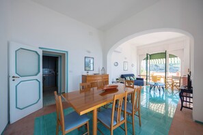 A dining area with a wooden table and chairs, a kitchen in the background, and a view through an archway to an outdoor patio.