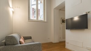 A modern living room with a grey sofa, a flat-screen TV mounted on the wall, and a window with a view of buildings outside. - Book Lovers@ArenaTheatre (Verona)