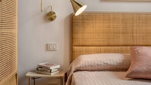 A neatly arranged bedroom with a woven headboard, a bedside table with books, and a wall-mounted lamp. - Book Lovers@ArenaTheatre (Verona)