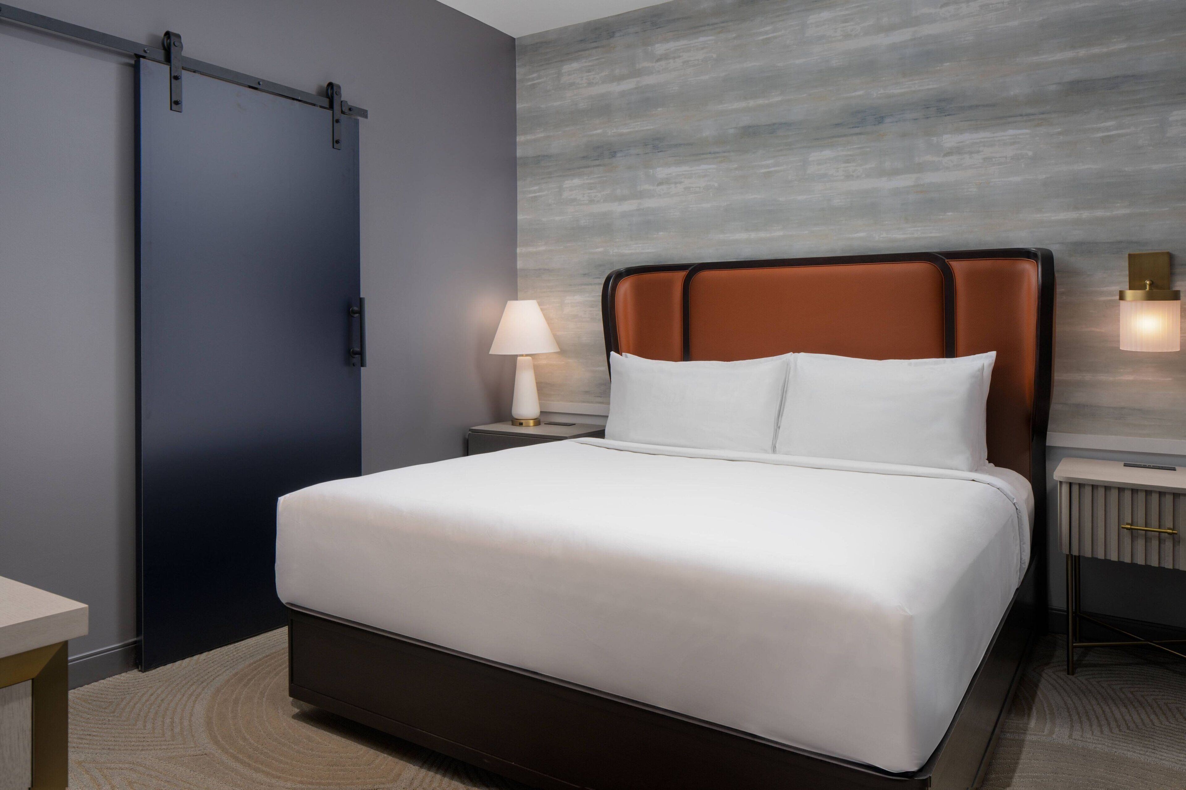 A neatly made bed with white linens, a brown headboard, and a bedside lamp.