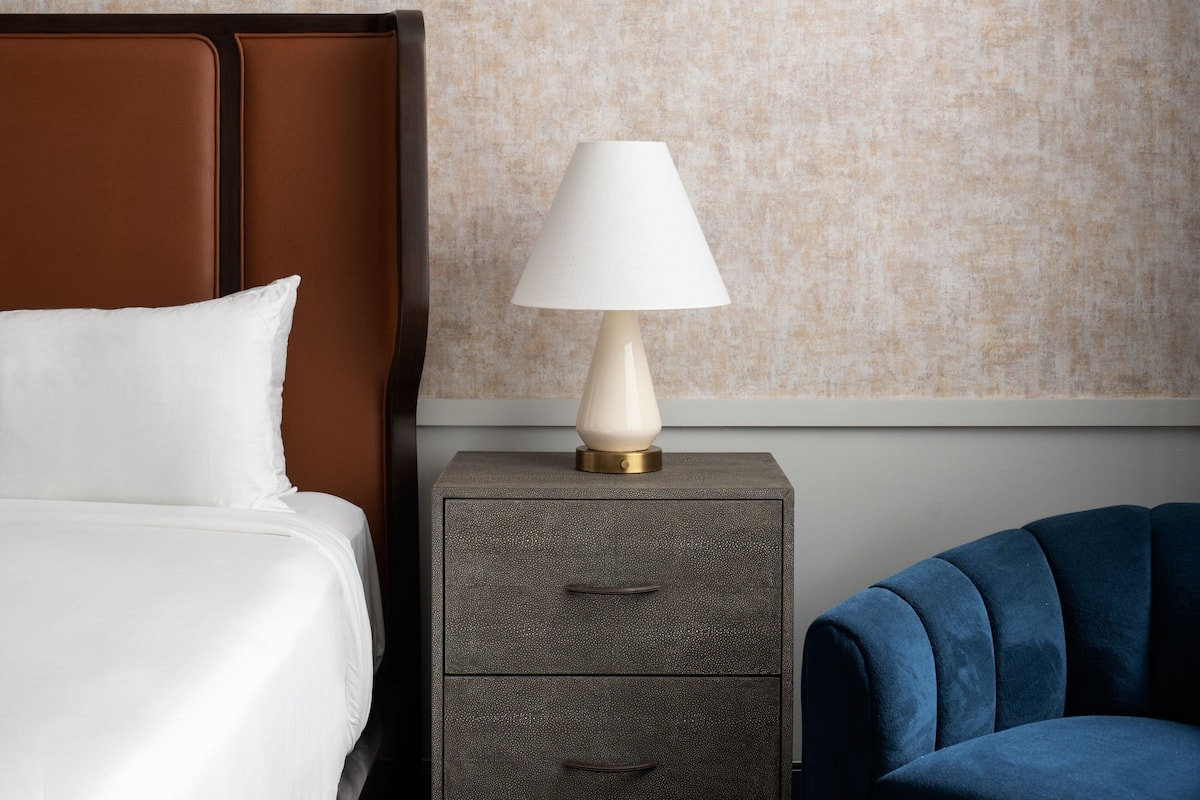 A neatly made bed with white linens, a bedside lamp on a wooden nightstand, and a blue upholstered chair.