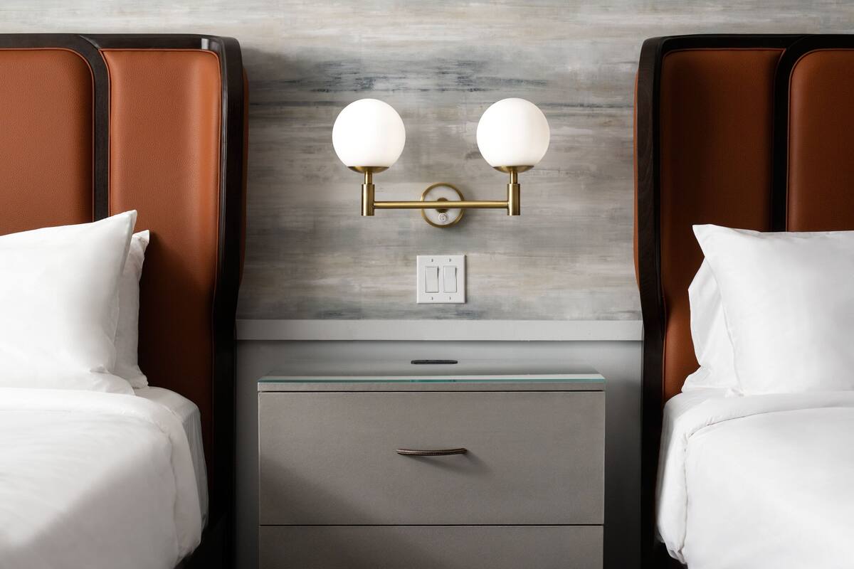 A hotel room with two beds, a nightstand with a drawer, a wall-mounted light fixture, and a power outlet.
