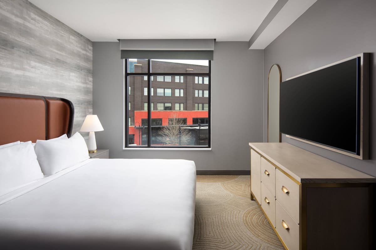 A hotel room with a large bed, a dresser with drawers, a flat-screen TV, a window with a view of buildings, and a lamp on the nightstand.