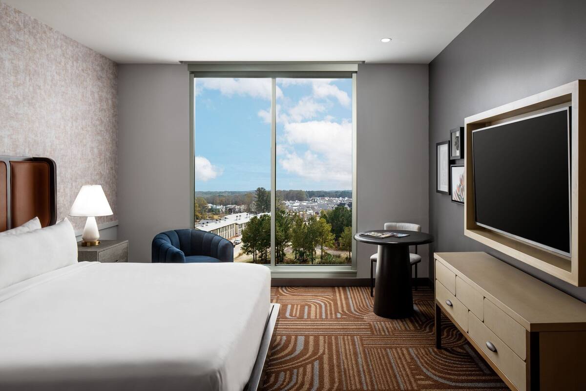 A hotel room with a large bed, a desk, a chair, a TV, and a view of the city.