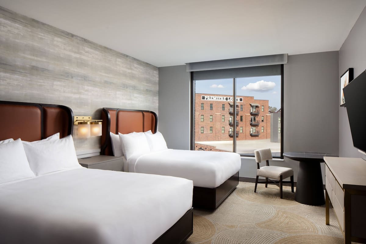 A hotel room with two beds, a desk, and a chair. There is a large window with a view of a building outside.