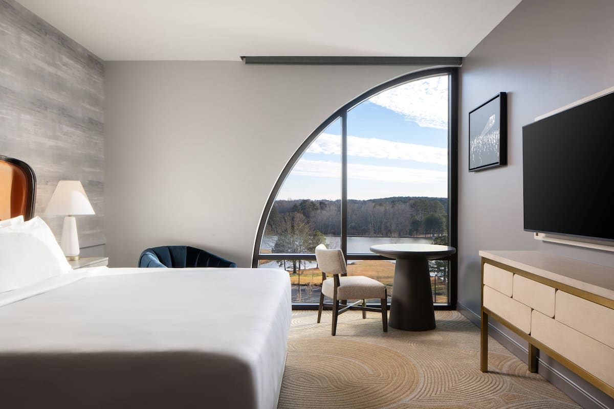 A modern hotel room with a large bed, a desk, a chair, a round table, and a TV.