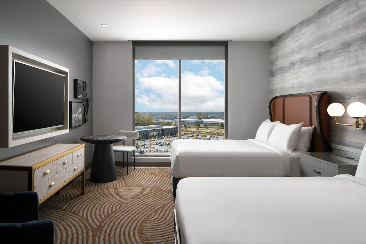 A modern hotel room with two beds, a large window with a city view, a flat-screen TV, and a small table.