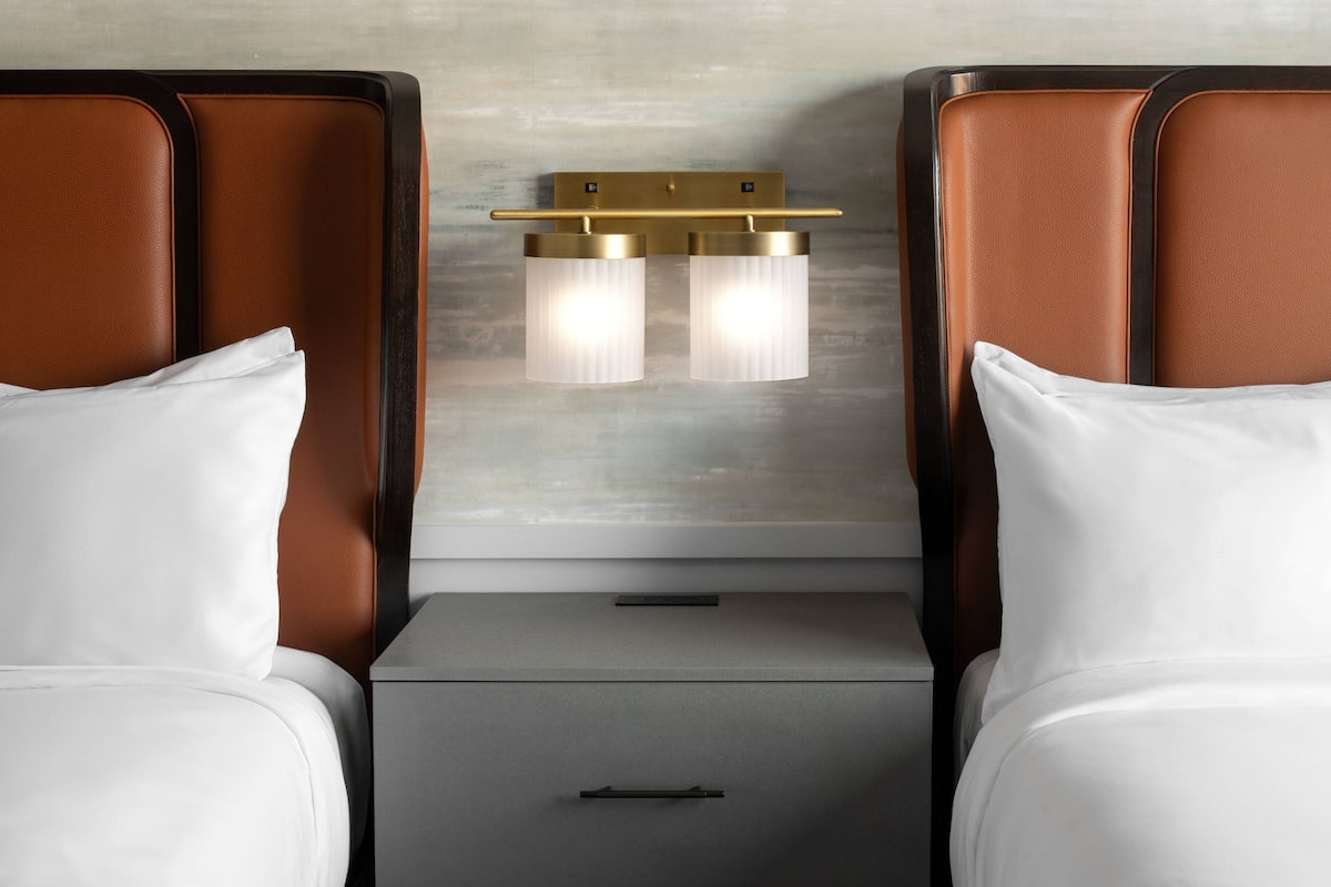 A hotel room with two beds, a nightstand, and a wall-mounted light fixture.
