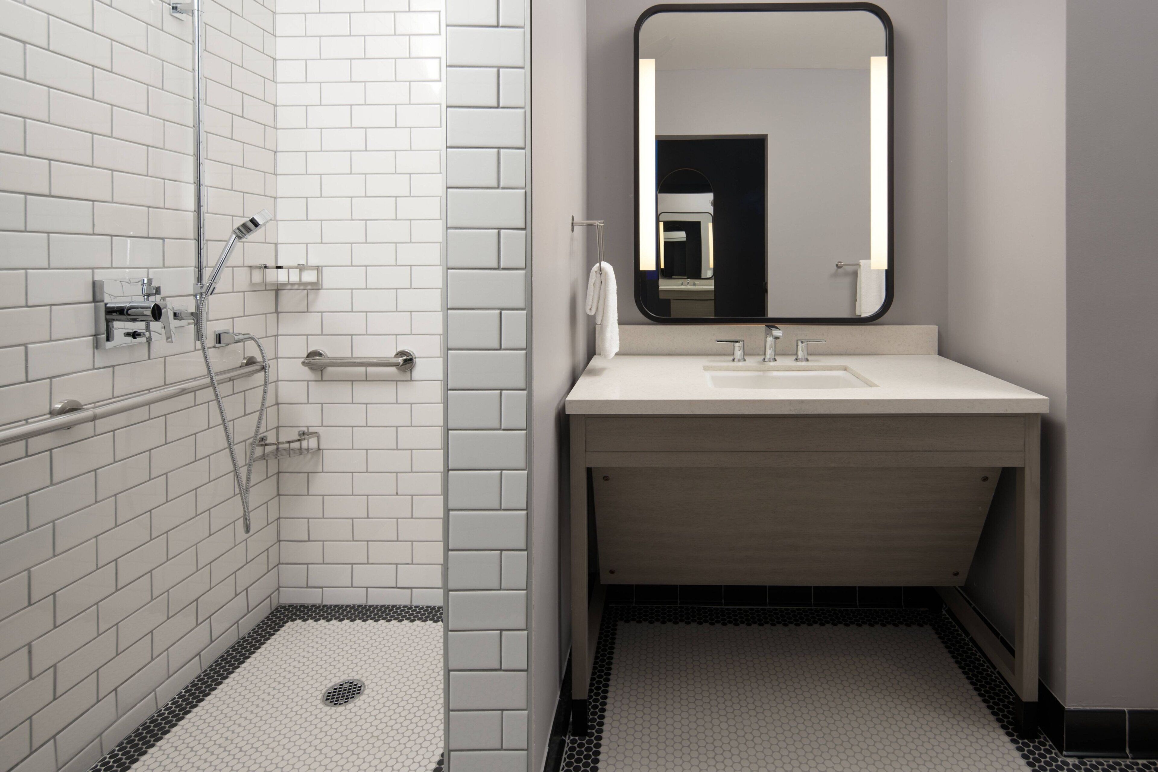 Un baño moderno con azulejos blancos tipo metro, una ducha con cabezal de ducha manual, un lavamanos con espejo y un perchero para toallas.