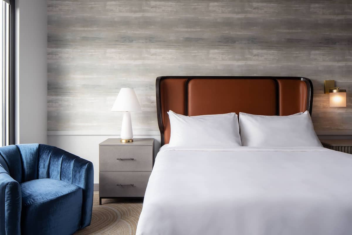 A modern hotel room with a large bed, a nightstand with a lamp, a chair, and a textured wall.