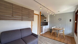 A compact living space with a sofa, a dining table with chairs, a kitchenette, and a clock on the wall.