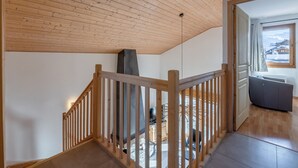 Una escalera de madera lleva a un área de estar con chimenea, un sofá y vistas de un paisaje nevado a través de una ventana.