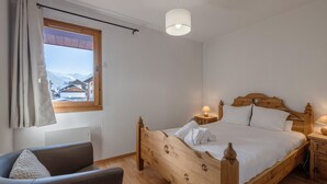 Un dormitorio con cama de madera, una silla, una mesita de noche, una lámpara y una ventana con vistas a edificios y montañas.