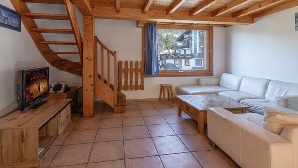 Una habitación acogedora con una escalera de madera, un sofá blanco, una mesita pequeña y un televisor sobre un mueble de madera.