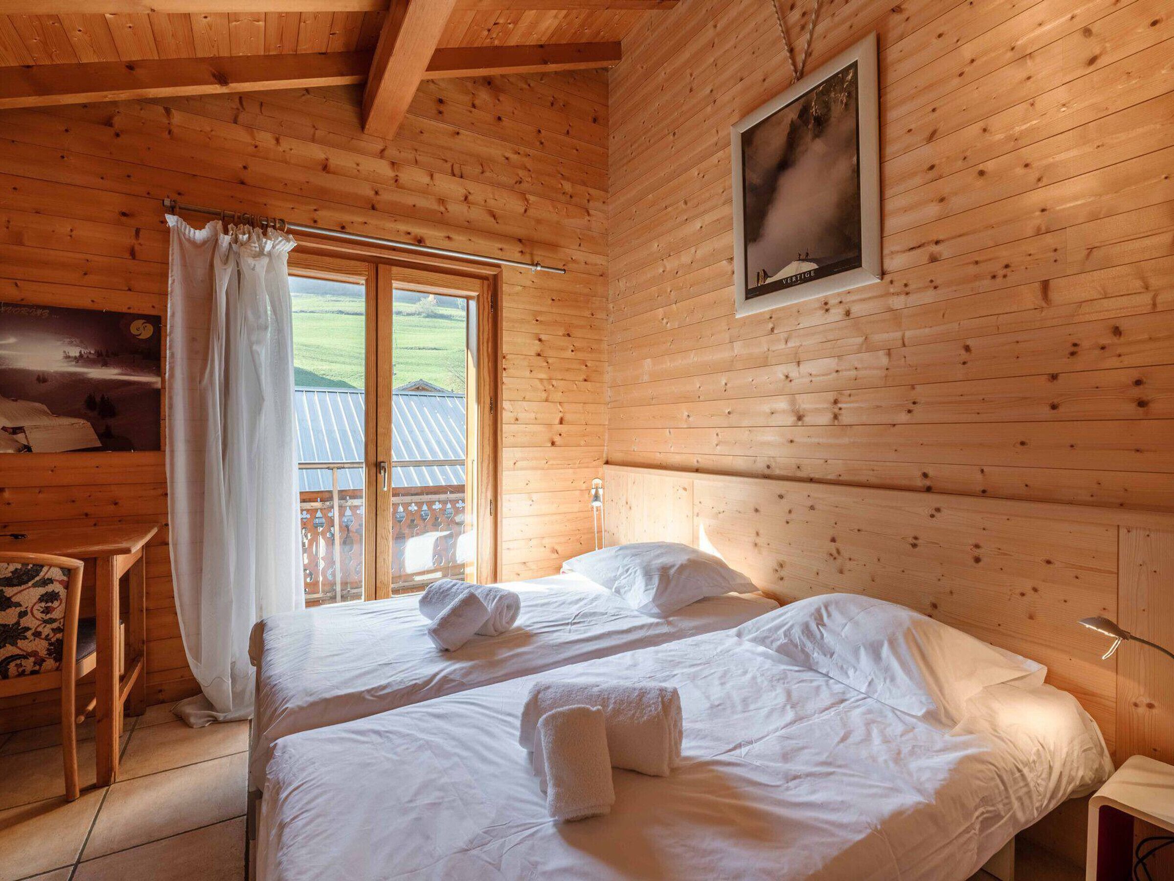 A wooden cabin room with two beds, a window with curtains, a bed with a pillow and a rolled towel, and a framed picture on the wall.