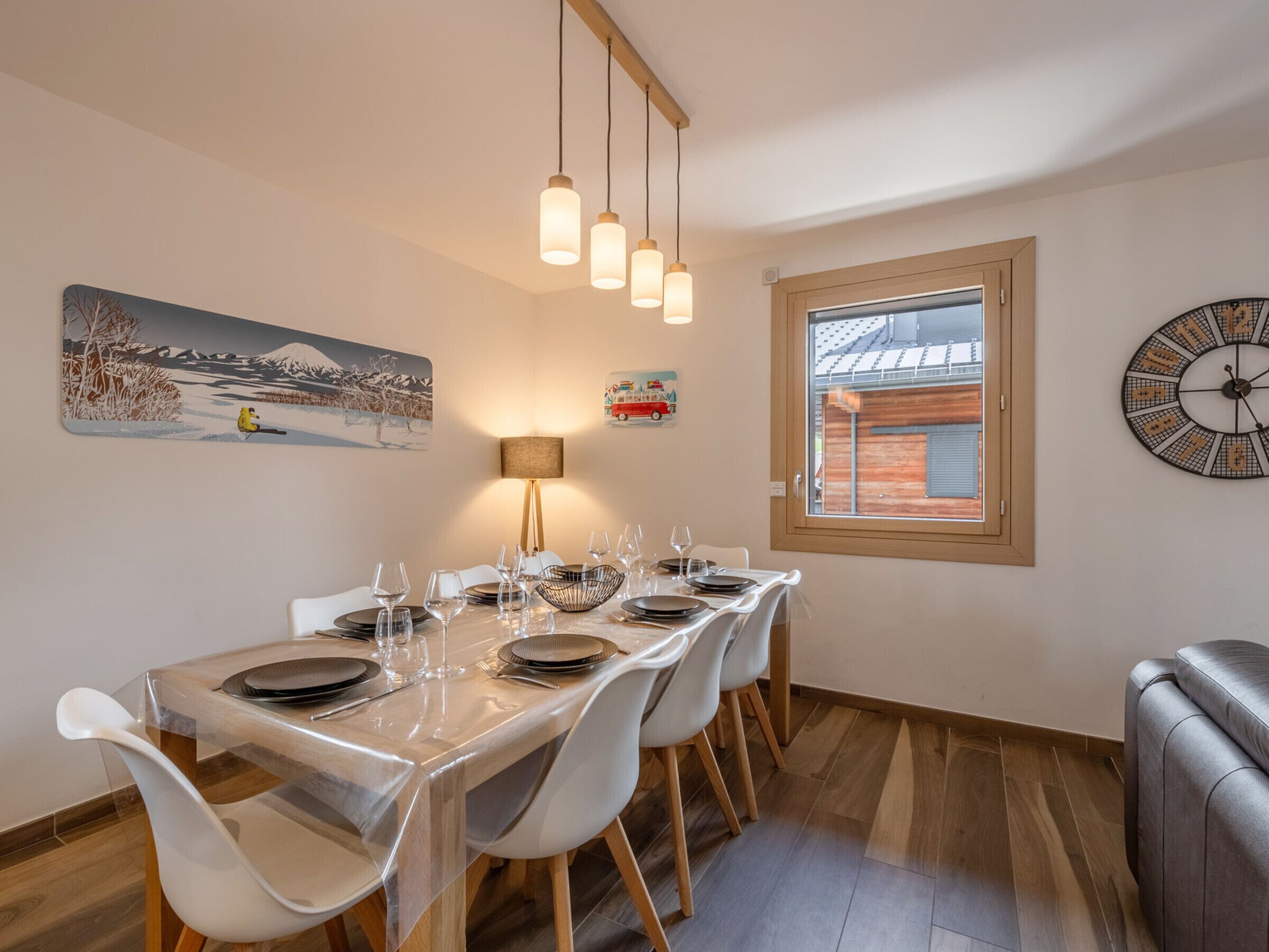 Een eetruimte met een tafel gedekt voor zes personen, een lamp, een klok en een raam met uitzicht op een gebouw.