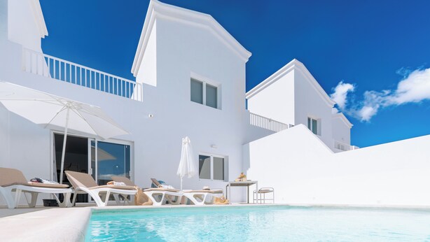 Une villa moderne de couleur blanche, dotée d’une piscine, de chaises longues et d’une petite table avec une chaise.