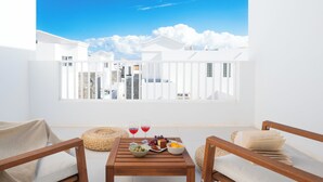 Un balcon d’où l’on peut admirer des bâtiments blancs et un ciel bleu dégagé. On y trouve une table en bois, sur laquelle sont posés un saladier rempli de fruits, un verre de vin rouge et un livre.