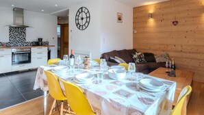 A dining area with a table set for four, a kitchen in the background, and a wooden wall with a clock.