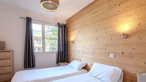 A bedroom with two beds, wooden walls, a window with curtains, and a bedside lamp.