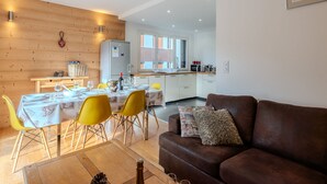 A modern living room with a brown leather sofa, yellow chairs, and a dining table set for four. A kitchen is visible in the background with white cabinets and a refrigerator.