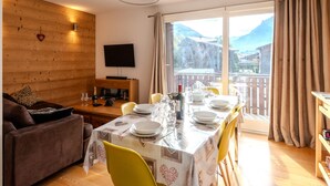 A dining area with a table set for four, a sofa, a TV mounted on the wall, and a view of mountains through the window.